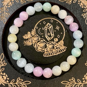 May include: A beaded bracelet featuring a variety of pastel-colored round gemstones. The bracelet is displayed on a black wooden coaster with a gold-colored floral and crystal design. The gemstones include shades of pink, blue, green, and cream.