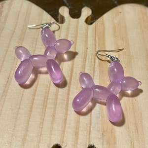 May include: A pair of lavender-colored balloon dog earrings. The earrings are made of translucent plastic and feature silver-colored ear wires. The earrings are displayed on a light-colored wooden surface.