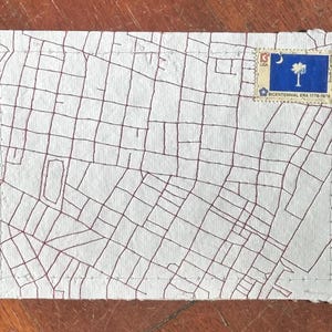 May include: A handmade, off-white envelope with a red grid pattern and a South Carolina Bicentennial stamp. The stamp features a blue background with a crescent moon and a palmetto tree. The envelope is on a wooden surface.