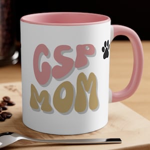 Puede incluir: Taza de café de cerámica blanca con borde y asa de color rosa. La taza tiene el texto "GSP MOM" en letras rosa y doradas con una huella de pata negra.