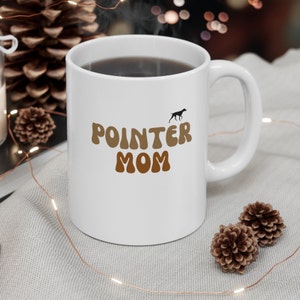 Puede incluir: Taza de café de cerámica blanca con un gráfico marrón y beige que dice "POINTER MOM" con una silueta de un perro caminando sobre el texto.