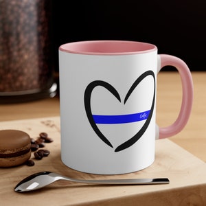 May include: White ceramic coffee mug with a pink rim and handle. The mug features a black hand-drawn heart with a blue line across the centre and the word "Cafe" written inside the heart.