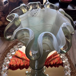 May include: A Murano glass vase with a ruffled rim and a flared base. The vase is a combination of clear, white, and pale green glass. The base has a starburst design. The vase has a label that says "Murano, Made in Italy."