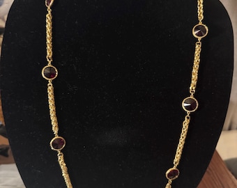 May include: A gold-toned necklace with a series of round, dark red gemstones. The necklace features a rope-like chain design with the gemstones evenly spaced along its length. The necklace is displayed on a black velvet surface.
