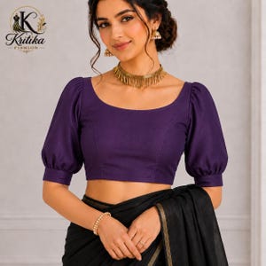 May include: A deep purple blouse featuring a wide scoop neckline and elbow-length puff sleeves with banded cuffs. The garment is paired with a black fabric draped over the lower torso. A gold-toned fringe necklace and gold-toned jhumka earrings.