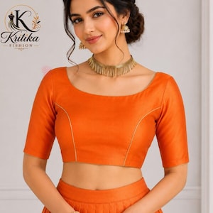 May include: A solid orange blouse featuring a wide scoop neckline, elbow-length sleeves, and thin gold piping accents along the front seams. The garment is paired with a matching orange skirt, a gold fringe choker necklace, and gold drop earrings.
