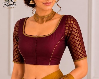 Può includere: Una blusa color bordeaux intenso con scollo quadrato e bordo dorato, maniche a gomito con un motivo geometrico dorato ripetuto e un corpetto bordeaux a tinta unita. Il capo è abbinato a un tessuto giallo senape drappeggiato in vita.