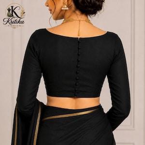 May include: A black long-sleeved blouse featuring a row of small fabric-covered buttons down the centre back. The garment has a wide neckline and is paired with a matching black fabric piece that has a thin gold border along the edge.