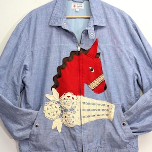 May include: A light blue zip-up jacket featuring a large red horse head appliqué with a dark brown mane and a cream-coloured lace floral detail positioned below the horse. The jacket has a collared neckline, long sleeves, and a front zipper closure.