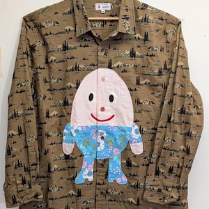 Peut inclure: Une chemise à manches longues boutonnée de couleur beige avec un imprimé répétitif de paysages de montagnes et de pins. Une grande applique en forme d'œuf avec un visage souriant, un haut rose et un bas à motif floral bleu est centrée.