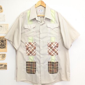 May include: Beige short-sleeve button-up shirt with light green and plaid accents. The shirt has two chest pockets and two lower pockets with plaid fabric. The collar and vertical stripes are light green.