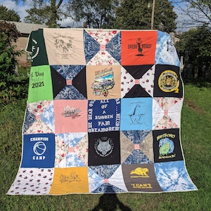 May include: A handmade quilt made from repurposed t-shirts. The quilt features a patchwork design with various graphic tees, including text and images. The color palette includes green, blue, red, and white. The quilt is displayed outdoors on a grassy area.
