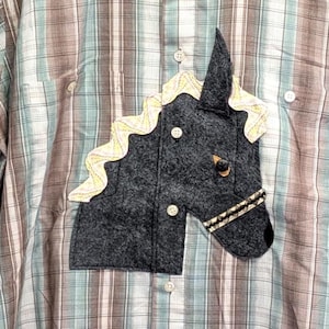May include: A short-sleeved button-down shirt featuring a plaid pattern in shades of brown, white, and light blue. A dark grey felt horse head appliqué with a cream-coloured mane and a decorative bridle is stitched onto the front of the garment.