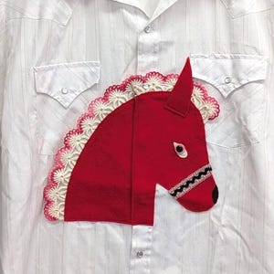 May include: A white short-sleeve button-down shirt featuring a red horse head appliqué on the chest. The horse design includes a white crocheted mane and a black and white bridle detail. The shirt has a collar, two chest pockets, and a button front.