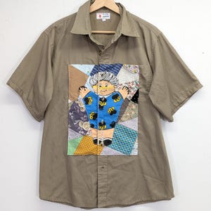 May include: Short-sleeved button-down shirt in a muted olive green. The shirt features a patchwork design on the front with a cartoon figure in a blue dress. The patchwork incorporates various fabric patterns and colors, adding a unique, handmade aesthetic.