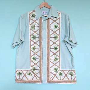 Peut inclure: Chemise vert clair à manches courtes avec une boutonnière et un col. La chemise présente un panneau décoratif avec des broderies de palmiers verts et marron, créant un thème tropical. La chemise est suspendue à un cintre en bois.
