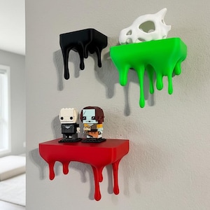 May include: Three wall-mounted shelves in black, green, and red, each with a dripping paint design. The shelves hold a white skull and two small figurines. The shelves are mounted on a grey wall.