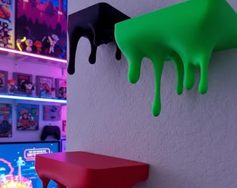 May include: Three wall-mounted shelves in black, green, and red, each with a unique dripping design. The shelves are mounted on a textured white wall, creating a modern and artistic display. Perfect for displaying collectibles or decor.