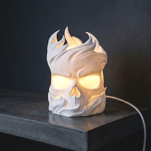 May include: A white skull-shaped lamp with stylised flame details surrounding the top and base. The eye sockets and top opening emit a warm, soft yellow glow. A thin white power cord extends from the side of the matte-finished object.