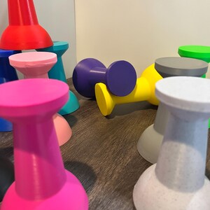 Pu&ograve; includere: Una collezione di piccoli oggetti a forma di rocchetto in vari colori solidi, tra cui rosa acceso, rosso, blu, giallo, viola, rosa chiaro, grigio e verde. Ogni oggetto presenta un'ampia base circolare e una parte superiore abbinata collegate da un collo stretto.