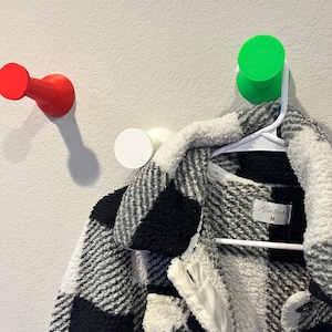 Pu&ograve; includere: Una giacca in sherpa a quadri bianchi e neri con etichetta del marchio Miss Posh taglia M, appesa a una gruccia di plastica bianca su un gancio da parete verde brillante. Altri due ganci da parete in rosso e bianco sono visibili sulla parete.