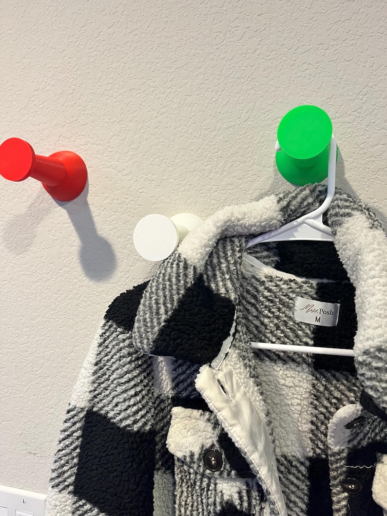 Pu&ograve; includere: Una giacca in sherpa a quadri bianchi e neri con etichetta del marchio Miss Posh taglia M, appesa a una gruccia di plastica bianca su un gancio da parete verde brillante. Altri due ganci da parete in rosso e bianco sono visibili sulla parete.