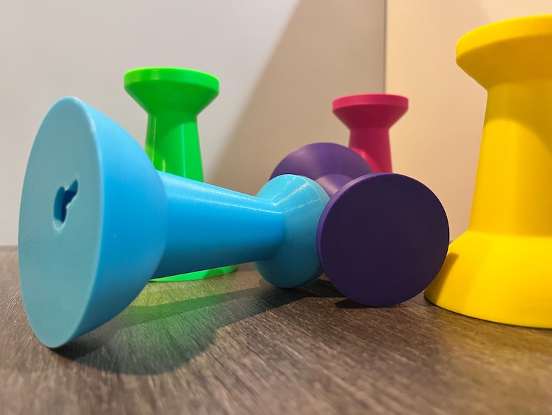 Pu&ograve; includere: Una collezione di cinque ganci da parete in plastica a forma di puntina da disegno in colori vivaci tra cui blu, verde, viola, rosa e giallo. Ogni gancio presenta un'ampia base circolare e uno stelo stretto, con un foro a serratura sul blu.