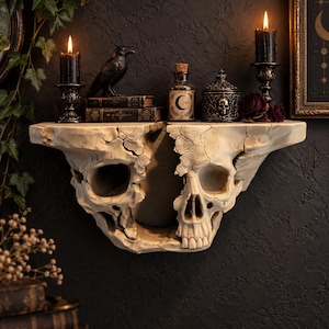 May include: A wall-mounted shelf shaped like a cracked, off-white human skull. The shelf holds two black candles in ornate holders, a black raven figurine, two stacked books, a glass bottle with a crescent moon label, and a lidded skull jar.