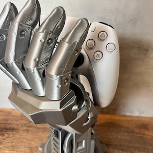 May include: A silver-coloured, robotic hand-shaped gaming controller stand. The stand holds a white gaming controller, displaying the controller's buttons. The base of the stand has a gear-like design, enhancing its mechanical aesthetic.