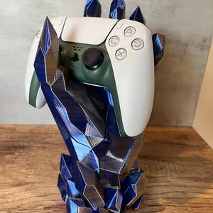 May include: A white and green video game controller is held by a blue crystal hand stand. The stand is made of 3D printed plastic and has a textured, metallic finish.