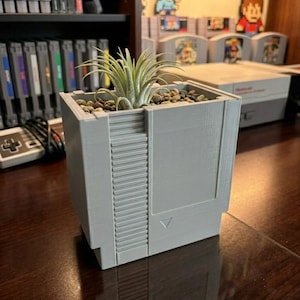 May include: A gray planter shaped like a classic video game cartridge, filled with small rocks and a green air plant. The planter has a textured design and is sitting on a wooden surface. The overall theme is a retro gaming aesthetic.