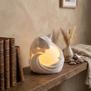 May include: A white cat-shaped lamp featuring a smooth, matte finish. The sculpture depicts a cat curled around a glowing, warm-toned spherical light source. The design incorporates fluid, curved lines that form the cat's body and tail, creating a soft, diffused illumination from within the center of the piece.