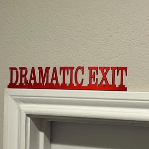 May include: A red metal sign that reads "DRAMATIC EXIT" is mounted above a white door frame. The sign is in a bold, sans-serif font and is a decorative element for a door or entryway.