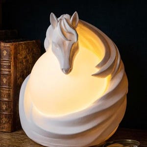 May include: A white sculptural table lamp featuring a horse head emerging from a swirling, rounded base. The lamp emits a warm, soft glow from behind the horse, illuminating the smooth, matte white surface of the structure in a circular pattern.