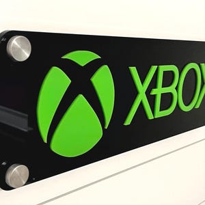 May include: A black and green Xbox logo sign with a 3D effect. The sign is mounted on a white wall with two silver screws.
