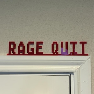 May include: A red, pixelated sign that reads "RAGE QUIT" is mounted above a white door frame. The sign is made of a solid material and is in a bold, blocky font, reminiscent of retro video games.