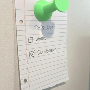 May include: A green push pin holds a lined paper to a white wall. The paper reads "TO-DO LIST" with checkboxes for "WORK" and "DO NOTHING", the latter checked. The paper has a torn edge.