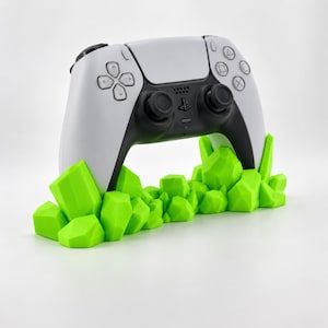 May include: A white and black wireless game controller resting in a bright neon green stand shaped like a cluster of jagged, geometric crystals. The controller features a central touchpad, two black analog sticks, and standard directional buttons.