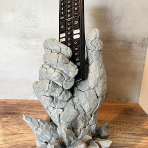 May include: A gray, stone-like hand holding a black remote control. The hand is standing on a base of gray, rocky crystals.
