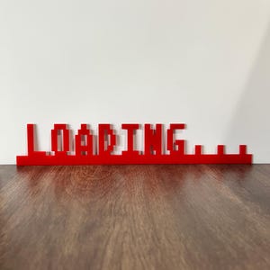 May include: Red pixelated "LOADING..." sign made of acrylic. The sign is on a wooden surface.
