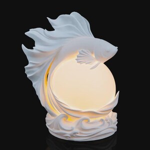 May include: A white sculptural lamp featuring a stylized betta fish with flowing fins positioned above a glowing, spherical light source. The base of the lamp is molded to resemble stylized ocean waves, providing a stable foundation for the illuminated orb.