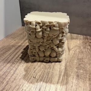 May include: A beige square-shaped box with a design of stacked skulls. The box is made of a smooth material and has a slightly textured surface.