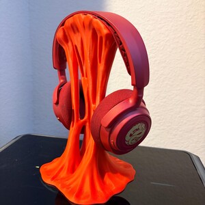Puede incluir: Auriculares rojos sobre un soporte naranja para auriculares. El soporte tiene un diseño abstracto y fluido. Los auriculares tienen almohadillas y diadema rojas. El soporte está sobre una superficie negra.