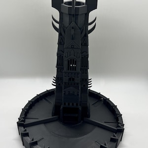 May include: A black 3D printed tower with a base, resembling a dark castle with pointed spires and a gate. The tower is on a circular base with a textured surface.
