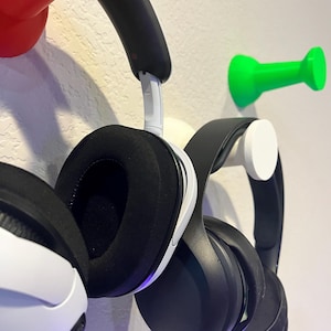 Pu&ograve; includere: Due paia di cuffie over-ear appese a ganci da parete. Un paio &egrave; bianco e nero con cuscinetti neri, l'altro &egrave; completamente nero. I ganci hanno la forma di grandi puntine da disegno nei colori rosso brillante, verde vivace e bianco.