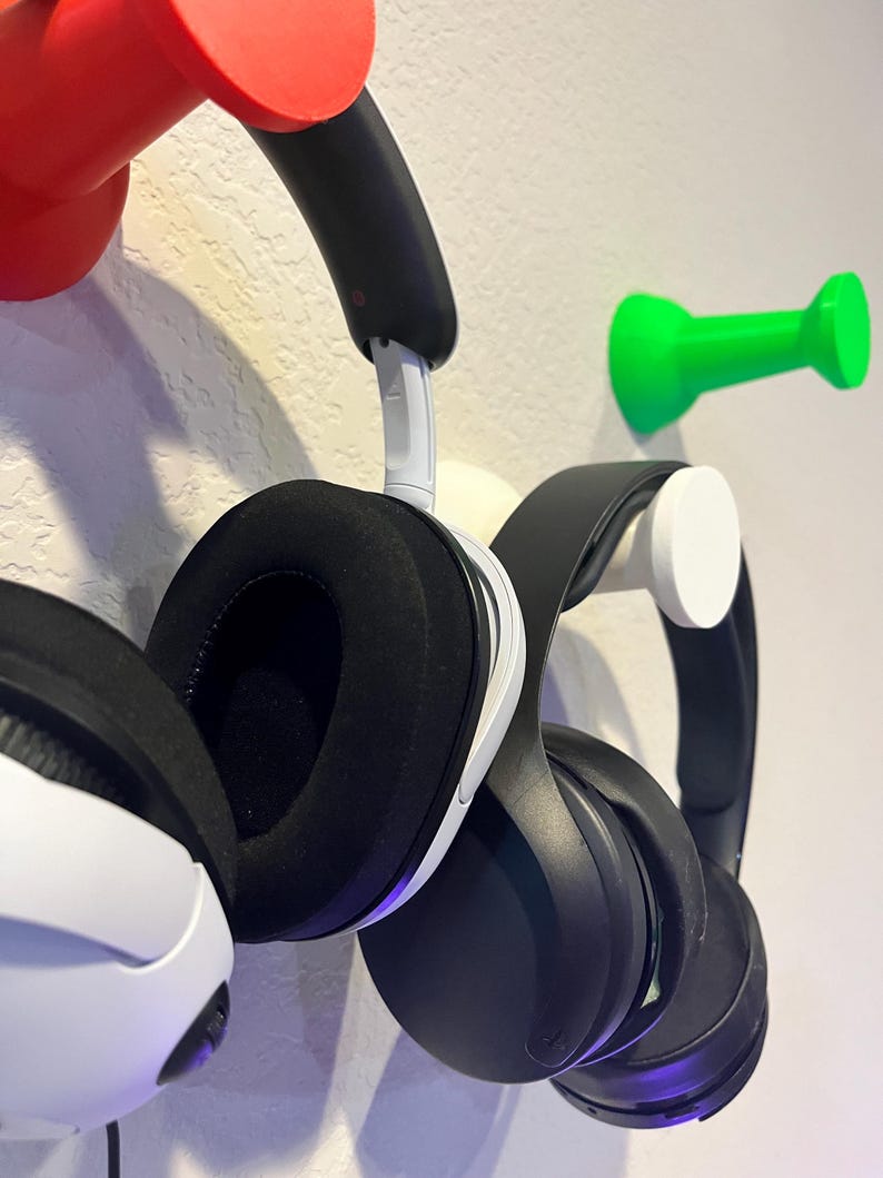 Pu&ograve; includere: Due paia di cuffie over-ear appese a ganci da parete. Un paio &egrave; bianco e nero con cuscinetti neri, l'altro &egrave; completamente nero. I ganci hanno la forma di grandi puntine da disegno nei colori rosso brillante, verde vivace e bianco.