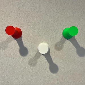 Pu&ograve; includere: Tre ganci da parete circolari disposti a forma di V. Il gancio a sinistra &egrave; rosso, quello a destra &egrave; verde brillante e il gancio centrale inferiore &egrave; bianco. Ogni gancio presenta una base cilindrica con una superficie piatta e circolare.