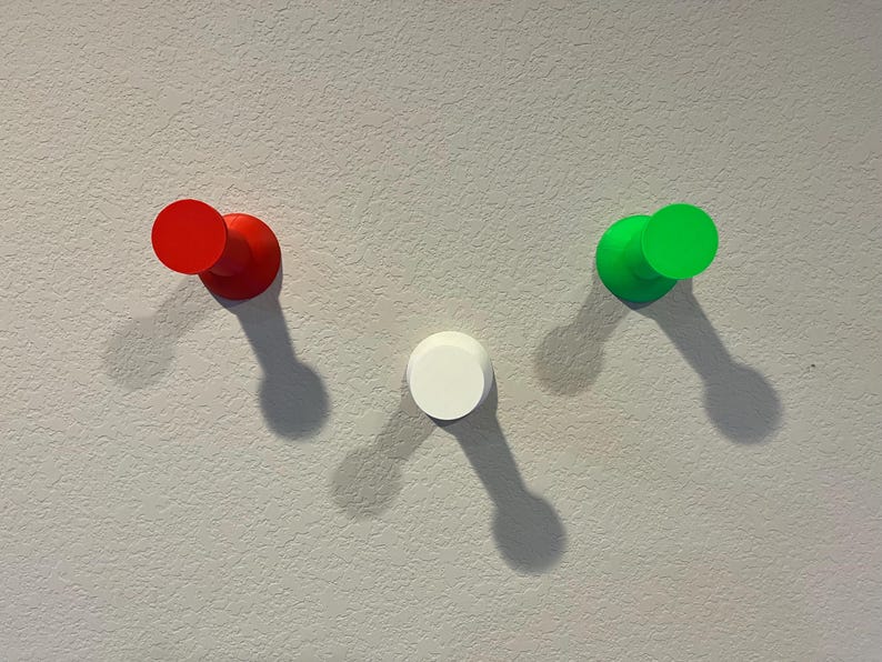 Pu&ograve; includere: Tre ganci da parete circolari disposti a forma di V. Il gancio a sinistra &egrave; rosso, quello a destra &egrave; verde brillante e il gancio centrale inferiore &egrave; bianco. Ogni gancio presenta una base cilindrica con una superficie piatta e circolare.