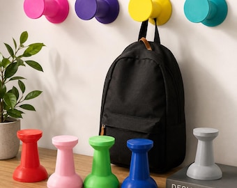 May include: A collection of colorful wall-mounted hooks and matching freestanding spool-shaped stands in pink, purple, yellow, teal, red, light pink, green, blue, and gray. A black backpack hangs from the yellow wall hook on the white wall.