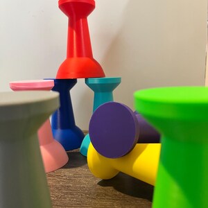 collection of colorful, abstract, giant pushpin-shaped objects in various colors, including red, blue, green, yellow, purple, and pink. The objects are arranged in a playful, stacked composition, with some lying on their sides.